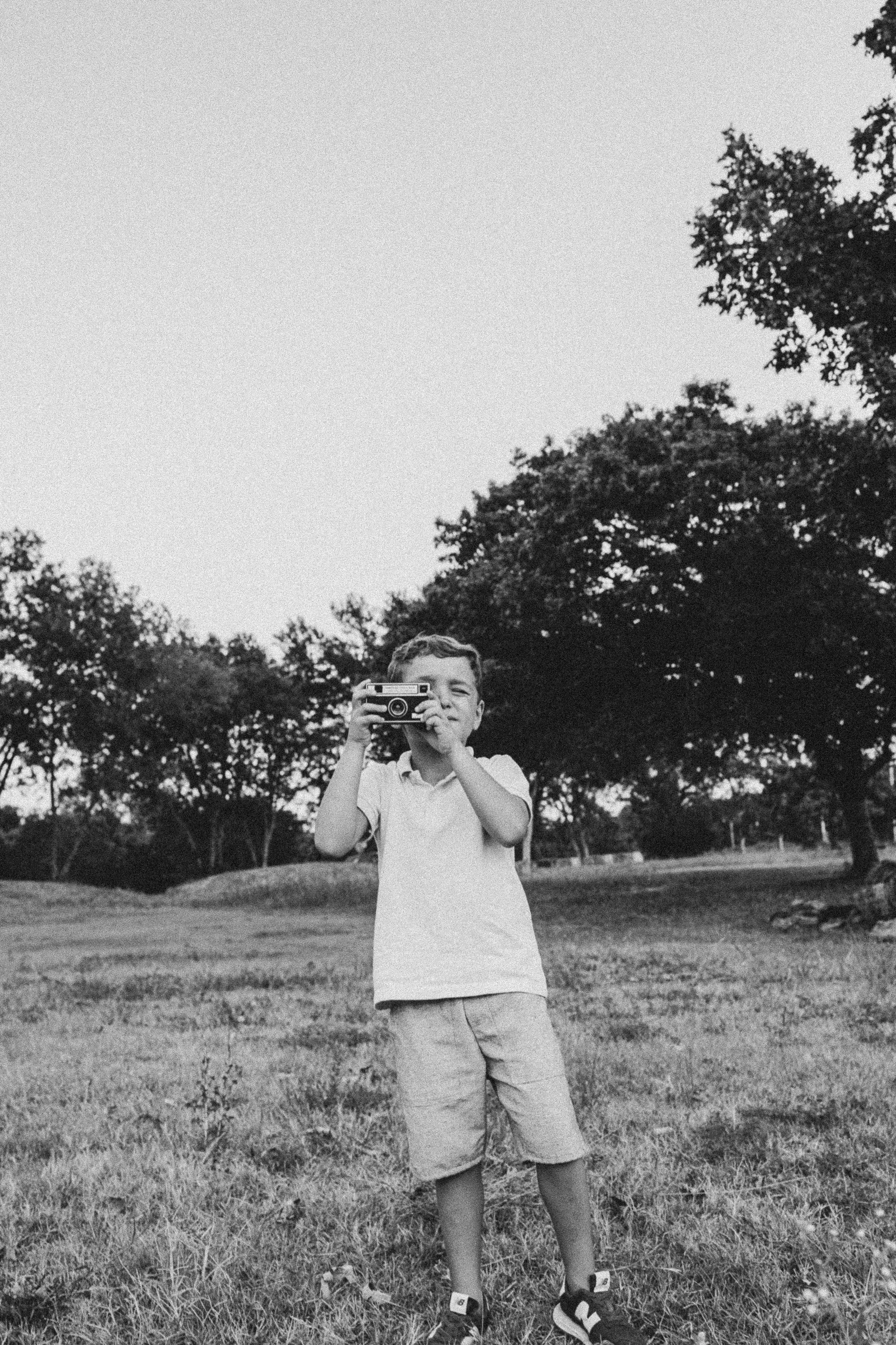 Boy in a polo shirt and shorts holding a film camera to his eye in a grassy field, black and white family session.