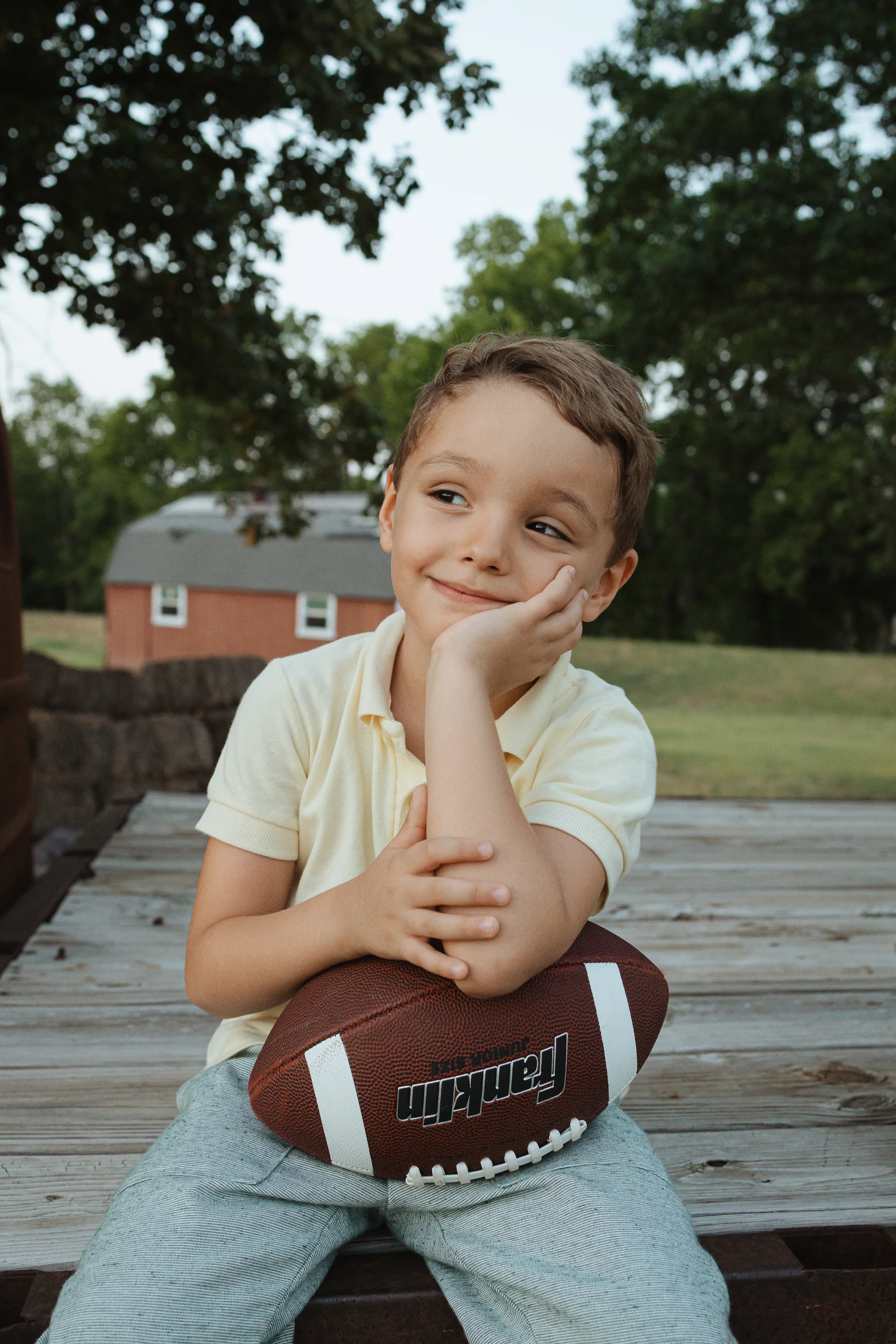 Young boy in a yellow polo resting his chin on his hand, holding a football on a wooden deck outdoors.