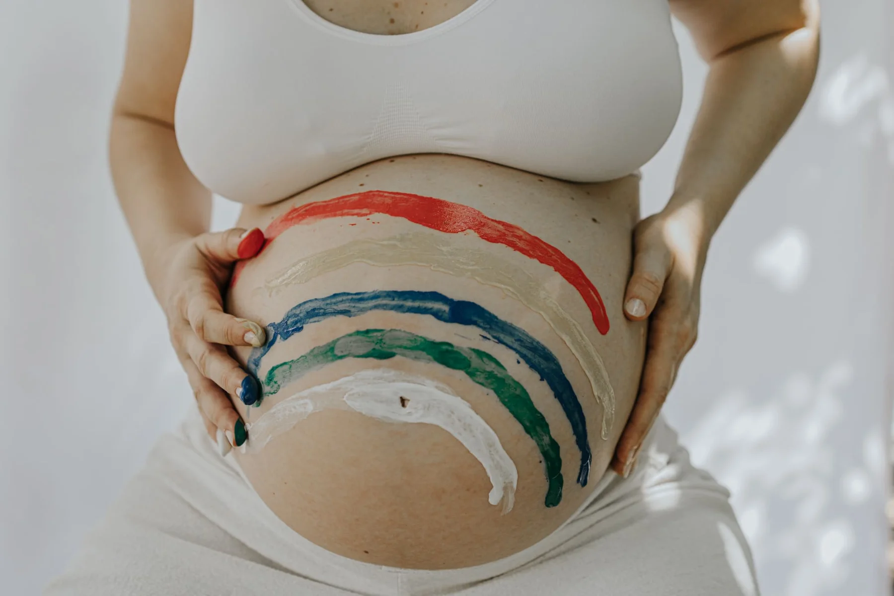 Pregnant woman cradling her belly painted with a rainbow, wearing a white top in soft light.